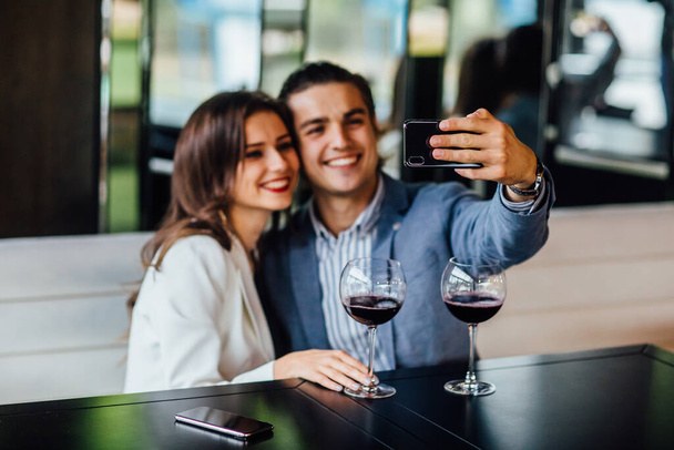 Cheerful loving couple is spending time together in modern restaurant. Attractive young woman in dress and handsome man in suit are having romantic dinner while make a selfie. Celebrating saint valentine's day. - Foto, imagen