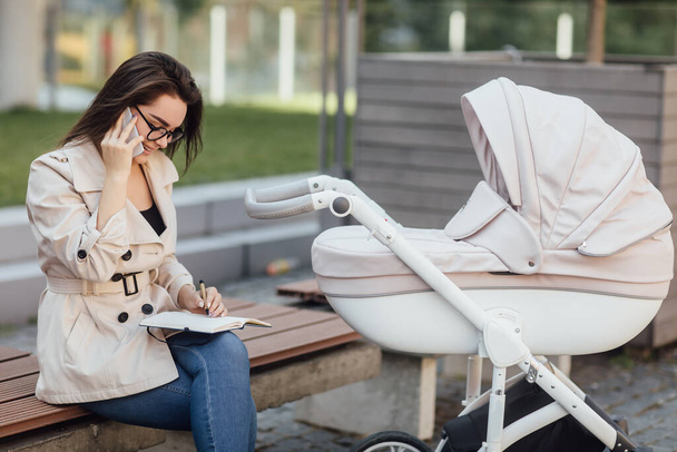 Happy mother sitting on the bench, holding phone and writing notes while her baby in stroller. - Foto, Imagem