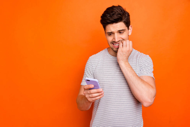 Oh no. Photo of terrified millennial guy hold telephone hand biting fingers read awful boss letter did bad thing wear striped t-shirt isolated bright orange color background - Foto, imagen