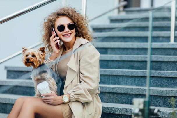 Business woman with curly hair in fashionable clothing sit on the city with her dog and speaking by phone. Lifestyle. - Foto, Imagem