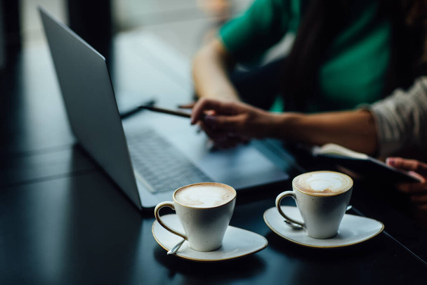Close up photo. Girls sitting in a restaurant at the table, chatting and using a laptop while have a break time. Business concept, latte on table. - 写真・画像