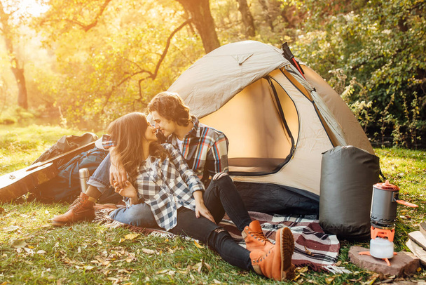 Young couple of tourists are exploring new places together. Happy beautiful woman and handsome man spending time together on nature, sitting near the tent on the blanket and hugging. - Photo, image