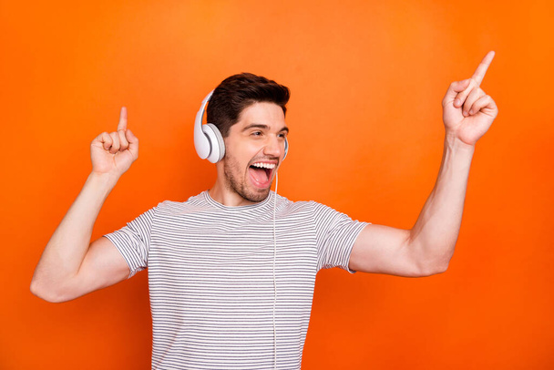 Photo of funky cool guy cheerful party mood chilling listening earphones direct fingers up empty space wear striped t-shirt isolated bright orange color background - Fotó, kép