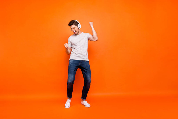 Full length photo of funky guy party mood chilling listen modern technology earphones raise hands dancing wear striped t-shirt jeans shoes isolated bright orange color background - Fotoğraf, Görsel