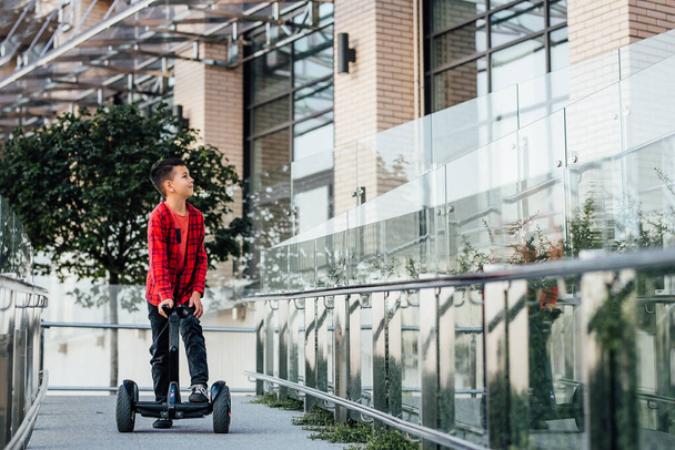 A boy with dark hair and red shirt rides a scooter. Modern schoolboy near the city building. Lifestyle. - Photo, image