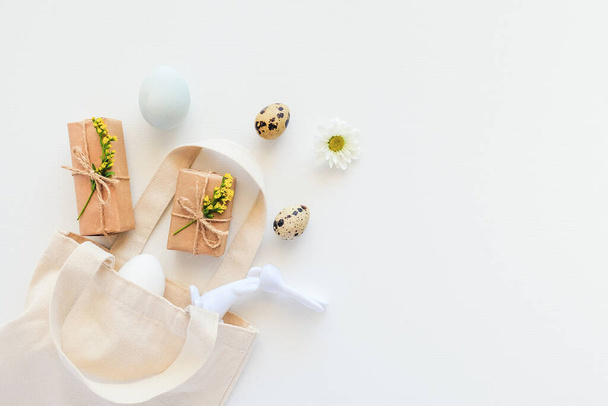 Happy easter concept. Eggs, yellow flowers, craft gift and bunny figurine fly out textile bag on white background Copy space Flat lay Top view. Template for greeting card, invitation, postcard. - Фото, зображення