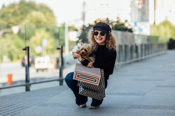 Full lenth of young woman with long curly hair holds small dog and shopping bags. Beautiful girl hugs little dog. Lady with puppy. Smiling attractive woman with Yorkshire terrier. Girl with dog in hands, and sale concept. - Photo, Image