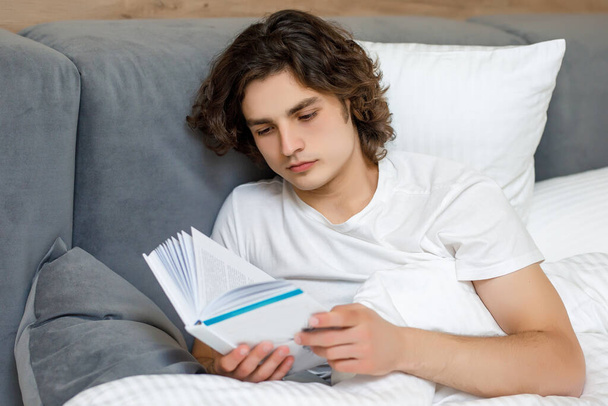 Красивий молодий чоловік лежить на ліжку читає книгу, відпочиває у своїй спальні
. - Фото, зображення