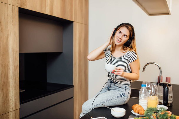 Blonde young woman in modern kitchen. Pretty woman having healthy breakfast while listening music by headphones. - Fotoğraf, Görsel