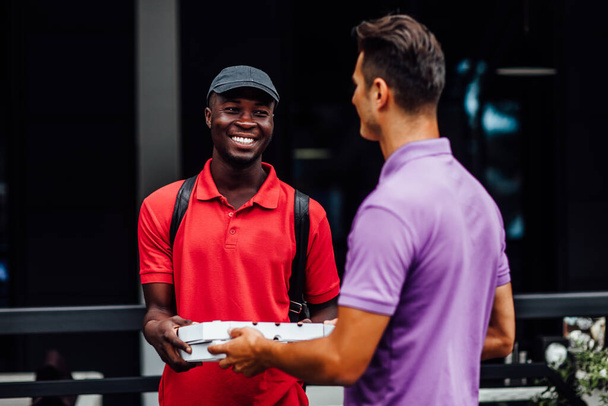 Mosolygós férfi alkalmazott pizzásdobozt vesz el egy fiatal afro-amerikai futártól, ételt rendelt a vállalati ebédszünetben. . - Fotó, kép