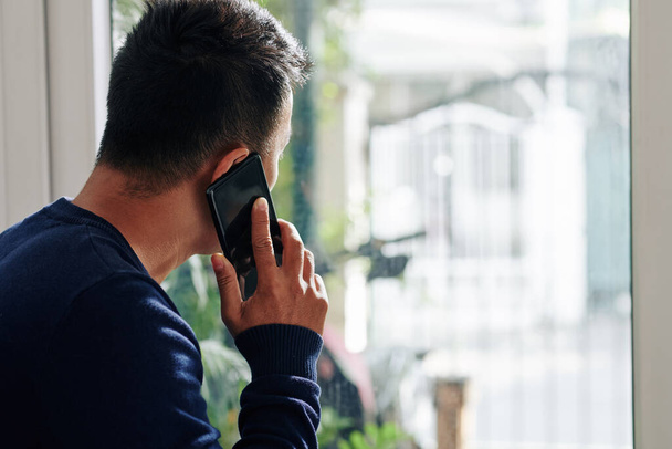 Jeune homme regardant par la fenêtre en parlant au téléphone avec un ami ou un collègue
 - Photo, image