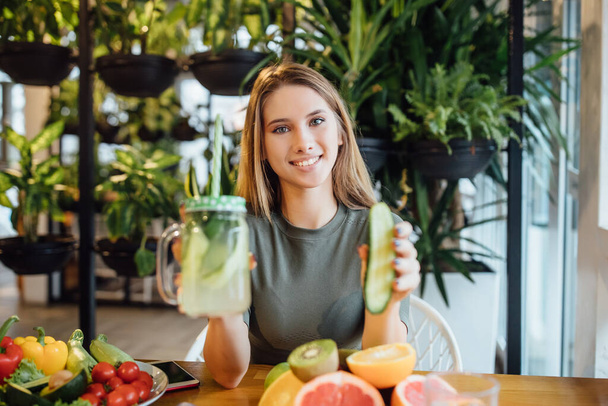 Photo of blonde fithess girl, trainer sit on the table and holding fresh cucumber smoothie, diet time concept. - Photo, Image