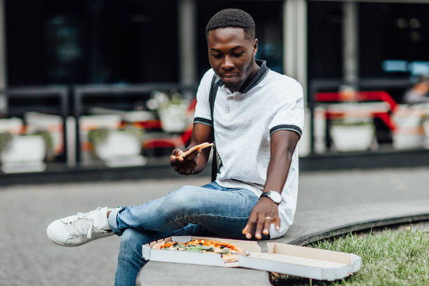 Guy dressed in a white T-shirt holds in his hands a piece of pizza and eats fast food on street. Young man eats a piece of pizza with his eyes closed. - Foto, afbeelding