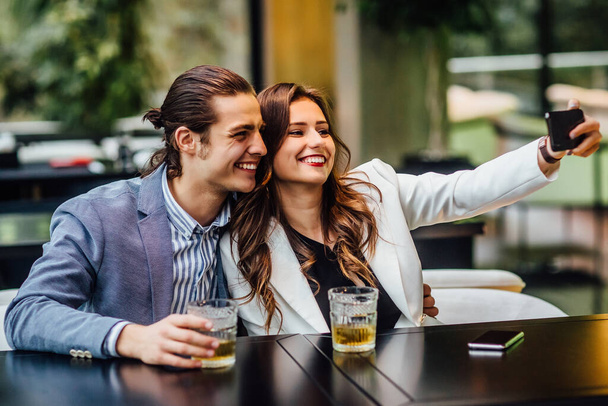 Lovely concept, pretty young and cheerful couple with drinks on table spend time at modern restaurant and make a selfie. - Photo, Image