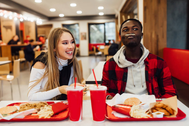 Beautiful couple having fun during lunch at fast food bar, sitting at table with drinks and food. - 写真・画像