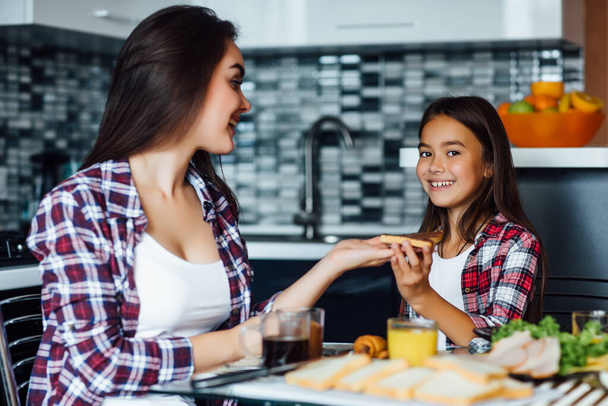 Happy girls in the kitchen. Mother and pretty child daughter are having breakfast with sandwich. - Zdjęcie, obraz