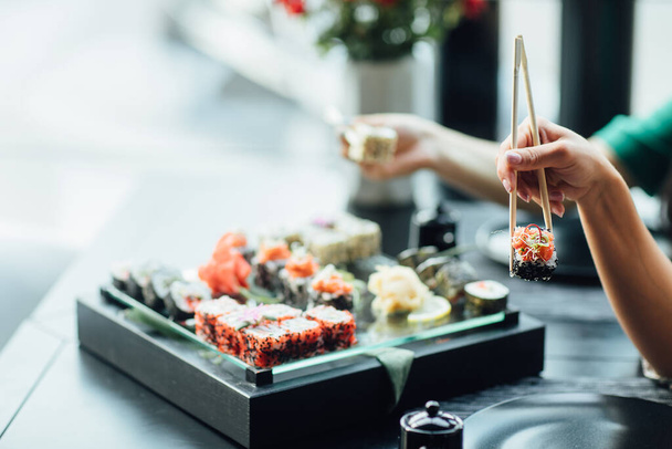 Chopstick holding sushi rolls set with salmon and cream cheese and cuccumber on black plate. Close Up. Uramaki, nori maki or futomaki sushi with trout fillets, soy sauce and wasabi. Sea food. - Fotoğraf, Görsel