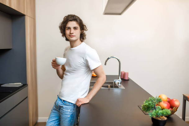 Portrait of handsome male in casual white t-shirt drinking morning coffee in kitchen, healthy time. - Photo, image