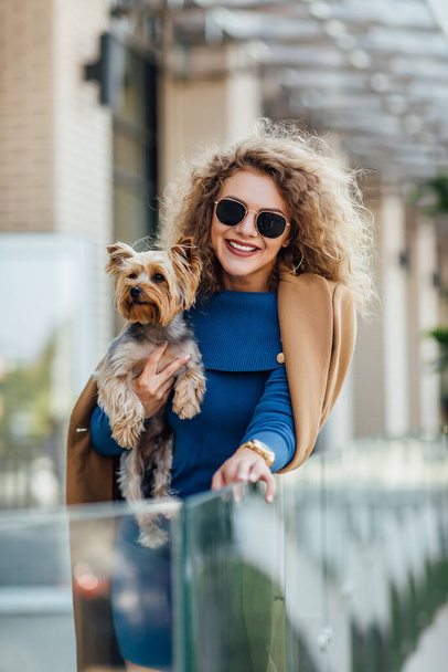 Successful attractive woman wearing at nude coat looking at camera and holding Yorkshire terrier. Yorkshire terrier concept. Fashion woman concept. - Фото, изображение
