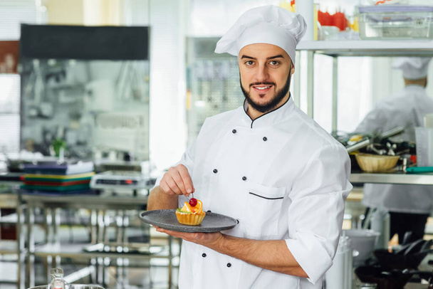 Portrait of the young, bearded chef in the kitchen of the restaurant with a ready-made sweet cake with fruits. - Fotoğraf, Görsel