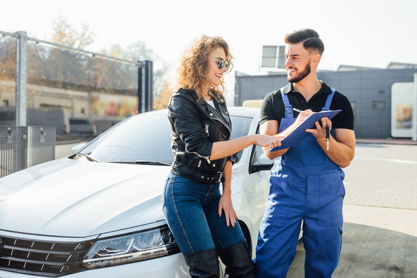 Blonde woman stands with a man who washes cars at the self-service, he holding blue folder and smile. - Photo, Image