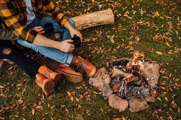 Close up photo of bonfire in the forest near tent. Happy couple drinks tea on the log near fire. - Φωτογραφία, εικόνα