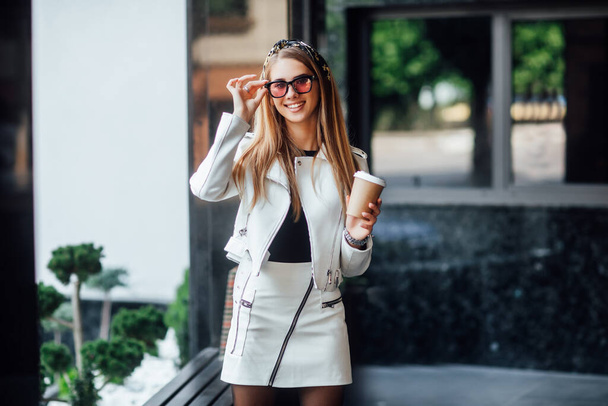 Portrait of young blonde model walking in the city, girl in stylish outfit drinking coffe and enjoying. - Zdjęcie, obraz