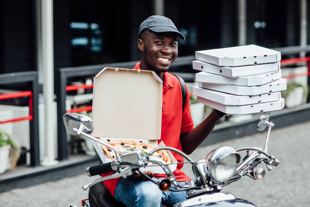 Fast food delivery concept. Happy busy courier or deliveryman shows fresh baked pizza on box, poses on fast scooter, delivers snack. Taste this delicious pizza. - Fotoğraf, Görsel