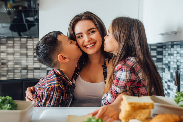 Young beautiful mother with her children that give a kissin mom, home kitchen during early breakfast. - Fotografie, Obrázek