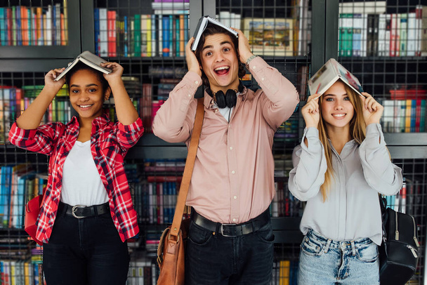 クラスメート、国際友情、楽しさと十代の概念。図書館でうろうろしている頭の上に屋根の形で本を持つカジュアルな服装で3人の陽気な10代の学生のグループ. - 写真・画像