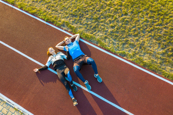 Top down aerial view of two young people sportsman and sportswoman laying on red rubber running track of a stadium field resting after jogging marathon in summer. - Foto, afbeelding