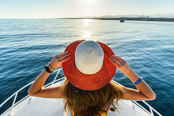 Junge Frau mit langen Haaren in gelbem Kleid und Strohhut steht auf weißem Jachtdeck und genießt Blick auf blaues Meerwasser. - Foto, Bild