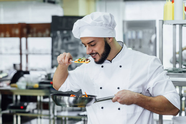 Ευτυχισμένος χαμογελαστός νεαρός σεφ με λευκή στολή ετοιμάζει ένα γεύμα με διάφορα λαχανικά. Υγιή και vegan τρόφιμα. - Φωτογραφία, εικόνα