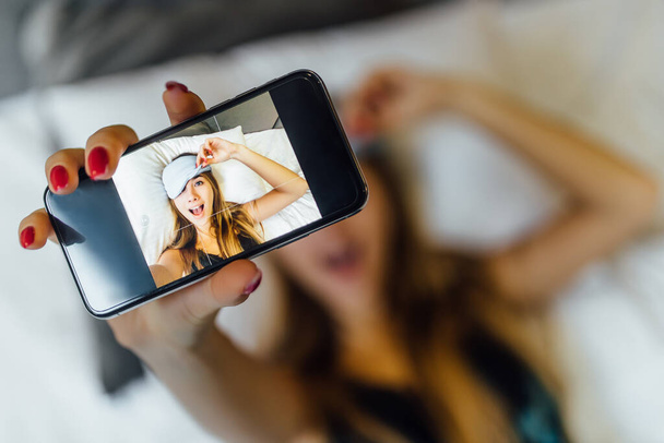 Portrait of young blonde woman lying on bed and making selfie by phone, wearing mask. - Foto, immagini