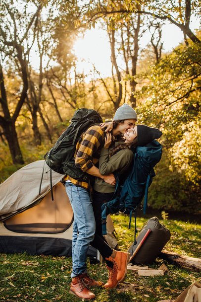Young beautiful couple with hiking backpack kissing in the forest near tent. Attractive woman and handsome man relaxing together in nature. Instagram stories picture. - Fotoğraf, Görsel
