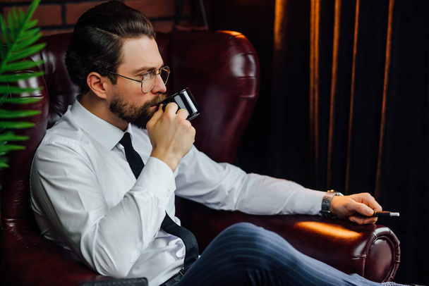 A man in a suit with a cup of coffee sits on a black leather sofa. - Zdjęcie, obraz
