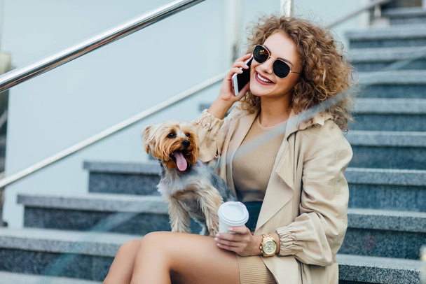 Прекрасная женщина с вьющимися волосами в модной одежде сидит в городе со своей собакой и разговаривает по телефону
. - Фото, изображение
