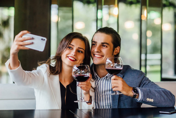 Beautiful loving couple is spending time together in modern restaurant. Attractive young woman in dress and handsome man in suit are having romantic dinner while make a selfie. Celebrating saint valentine's day. - 写真・画像