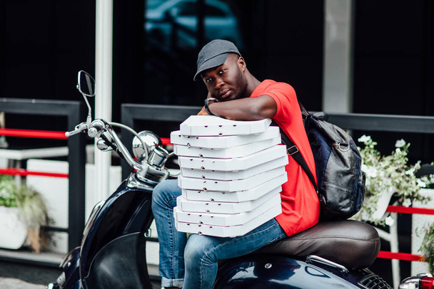 El tipo, ocupado con sus deberes, sostiene una pila de cajas de cartón, conduce scooter, usa casco y camiseta roja, trabaja en pizzería
. - Foto, Imagen