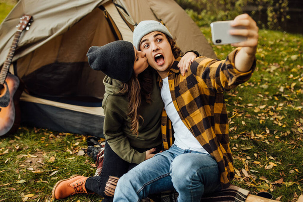 Jonge liefdevolle paar toeristen veel plezier en selfie maken in de buurt van het vuur in de natuur. Knappe man en mooie vrouw ontspannen in de buurt tent op de camping. - Foto, afbeelding