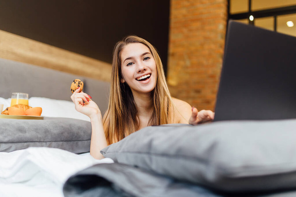 Photo of happy blonde woman using laptop on bedroom with cake in hand. - Foto, imagen