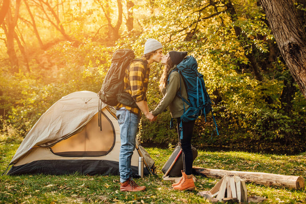 Young beautiful couple with hiking backpack kissing in the forest near tent. Attractive woman and handsome man relaxing together in nature. - Foto, Imagen