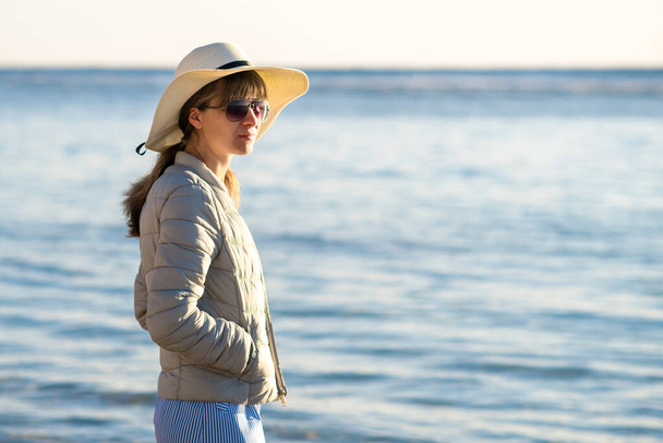 Young woman in straw hat and a dress standing alone on empty sand beach at sea shore. Lonely tourist girl looking at horizon over calm ocean surface on vacation trip. - Photo, Image