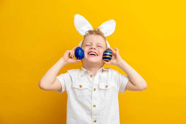 Cheerful boy in rabbit bunny ears on head on yellow studio background. Exited crazy smiling happy child. Easter blue color of the year painting eggs in hands. Easter and holidays - Фото, изображение