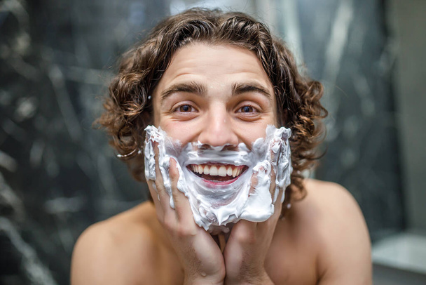 Young and happy man shaving with  shaving cream in bathroom, morning concept. - Фото, изображение