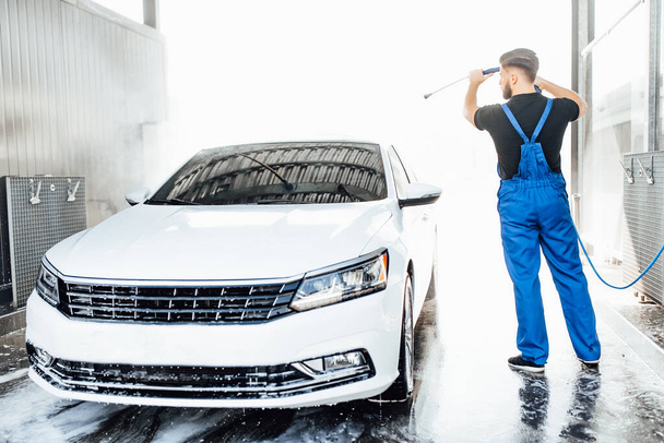 Professional young washer in blue uniform washing car with water gun on an open air car wash. - Foto, afbeelding