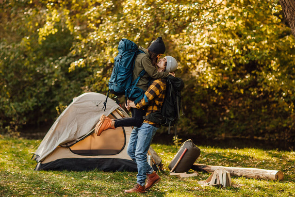 Young beautiful couple with hiking backpack kissing in the forest near tent. Attractive woman and handsome man relaxing together in nature. Instagram mood stories picture. Hugging people during trekking. - Fotoğraf, Görsel