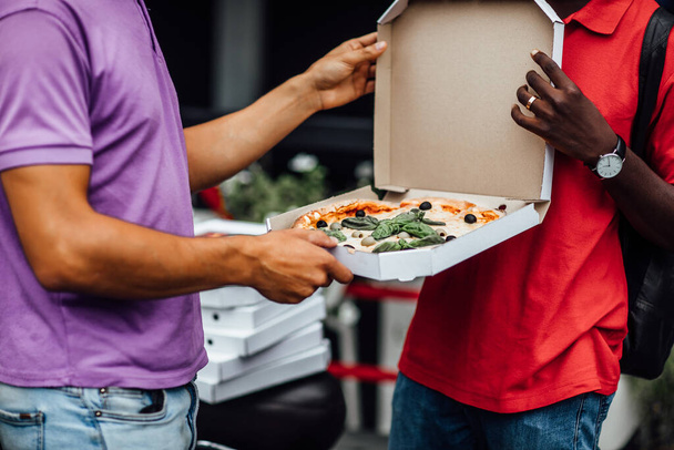 Close up photo. Courier delivery man in red uniform giving box of hot delicious pizza to customer, try it now. Candid emotion. - Fotografie, Obrázek