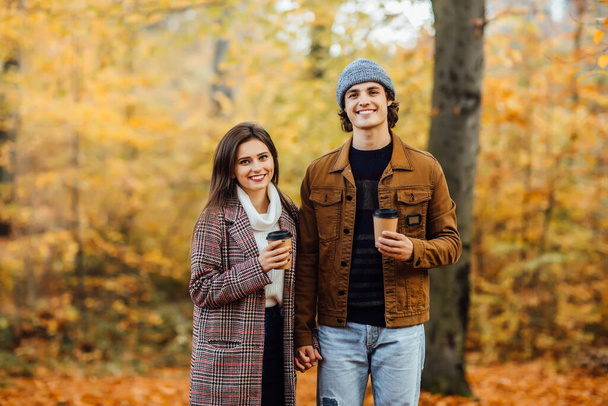 Family and hot autumn drink concept. Couple in love with scarves sits in park holding cups of tea or coffee. - Foto, Imagen