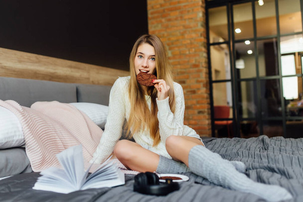 Relaxed playful woman wearing t-shirt, shorts and socks having lazy morning, lying in bed, having breakfast. People, leisure and spare time concept. - Фото, изображение